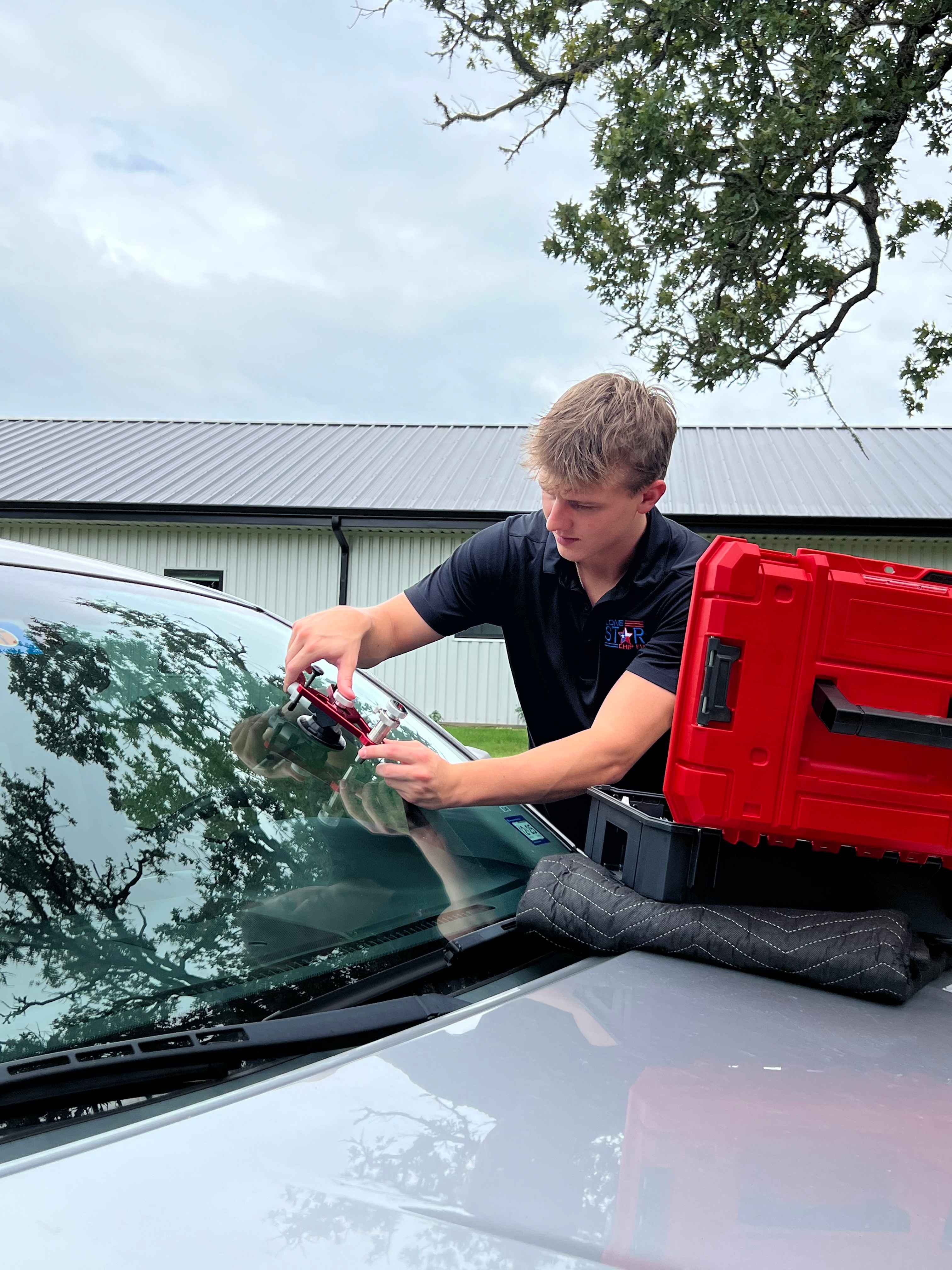 Young man repairing a car windshield with suction tools, focused and attentive. He wears a black shirt; a red toolbox is nearby on the hood. Overcast sky and trees in the background.