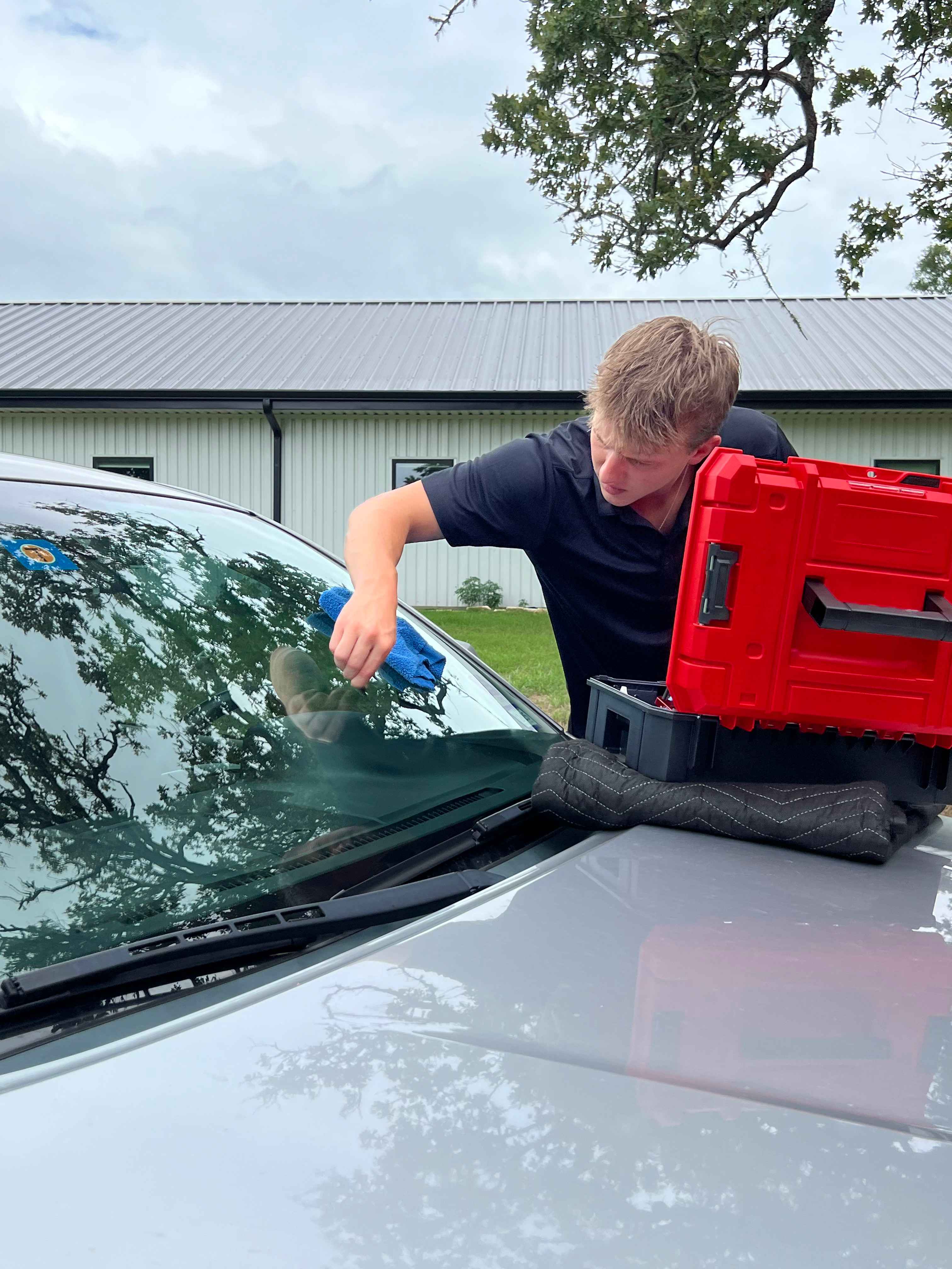 Young man repairing a car windshield with suction tools, focused and attentive. He wears a black shirt; a red toolbox is nearby on the hood. Overcast sky and trees in the background.